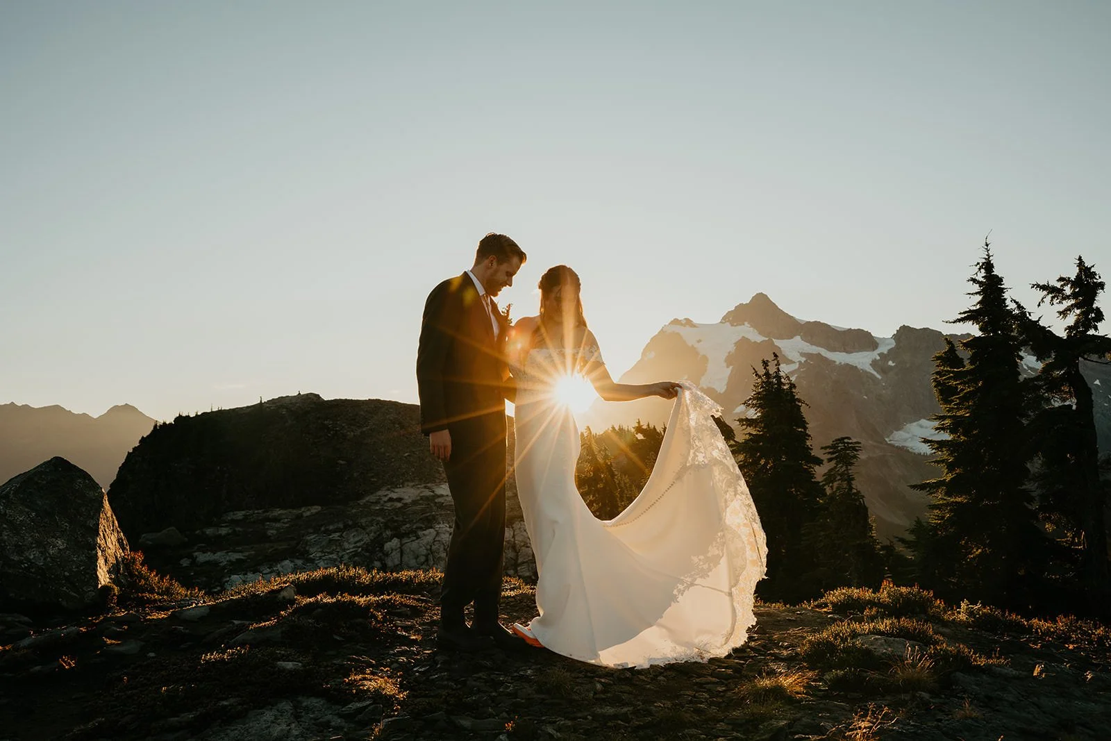 Couple holding hands on an alpine ridge in Washington as the sun sets behind Mount Rainier, golden light shining through the bride’s flowing dress during their mountain elopement.