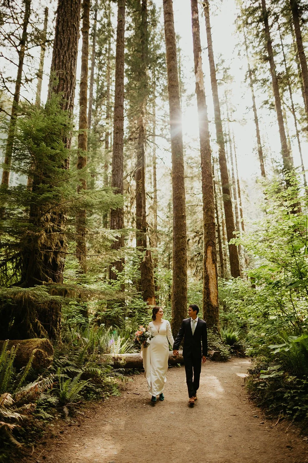 Couple walking hand in hand along a forest trail, towering trees and soft light surrounding them on all sides.