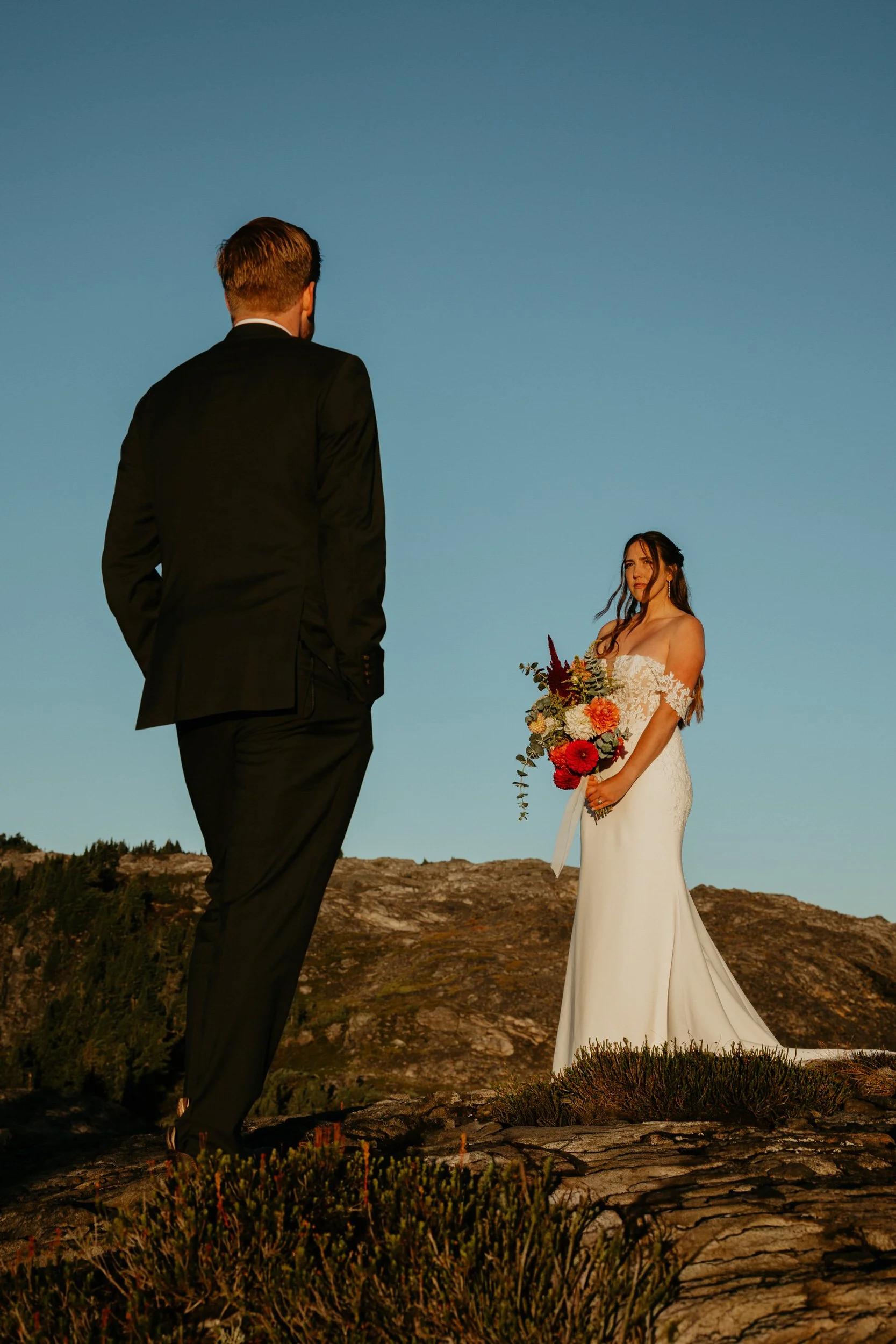 Bride and groom look at each other on a mountain ridge as the sun rises on them