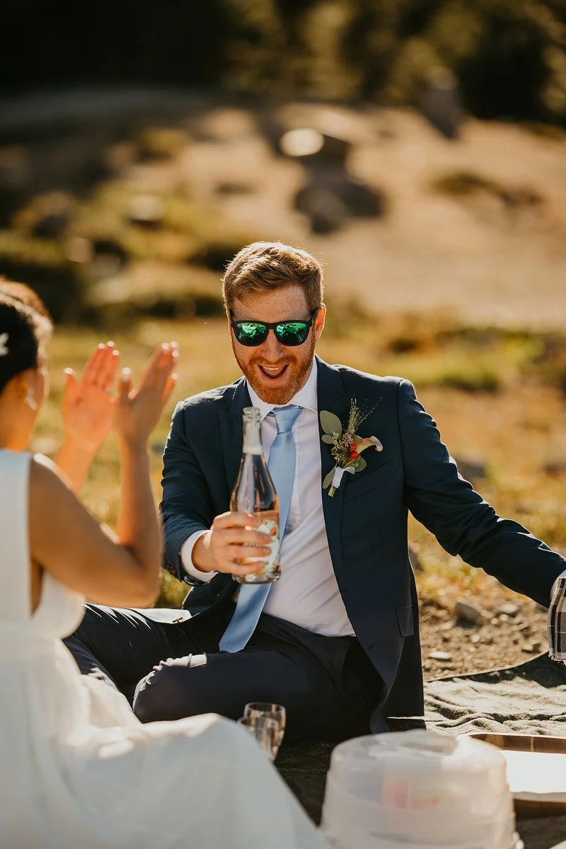 Groom pouring champagne while sitting on a blanket during a mountaintop elopement picnic in Washington.