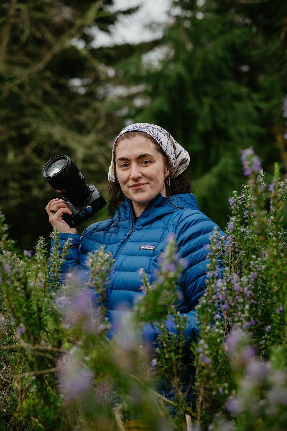 Washington elopement photographer smiling in a blue jacket while standing among lush evergreen trees.