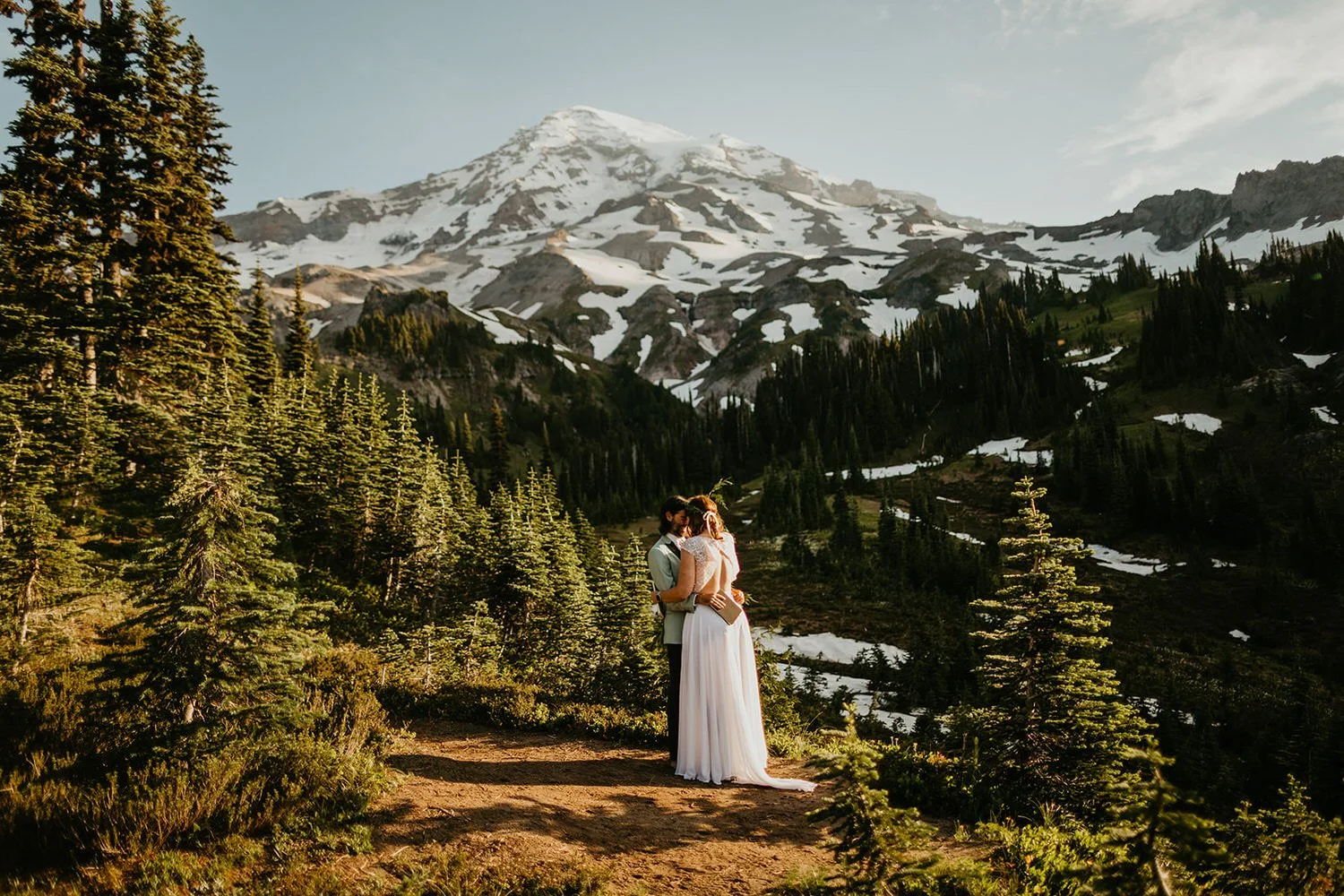 A couple embraces on a sunlit alpine overlook surrounded by evergreen trees, with Mount Rainier’s snow-covered slopes rising dramatically in the ba