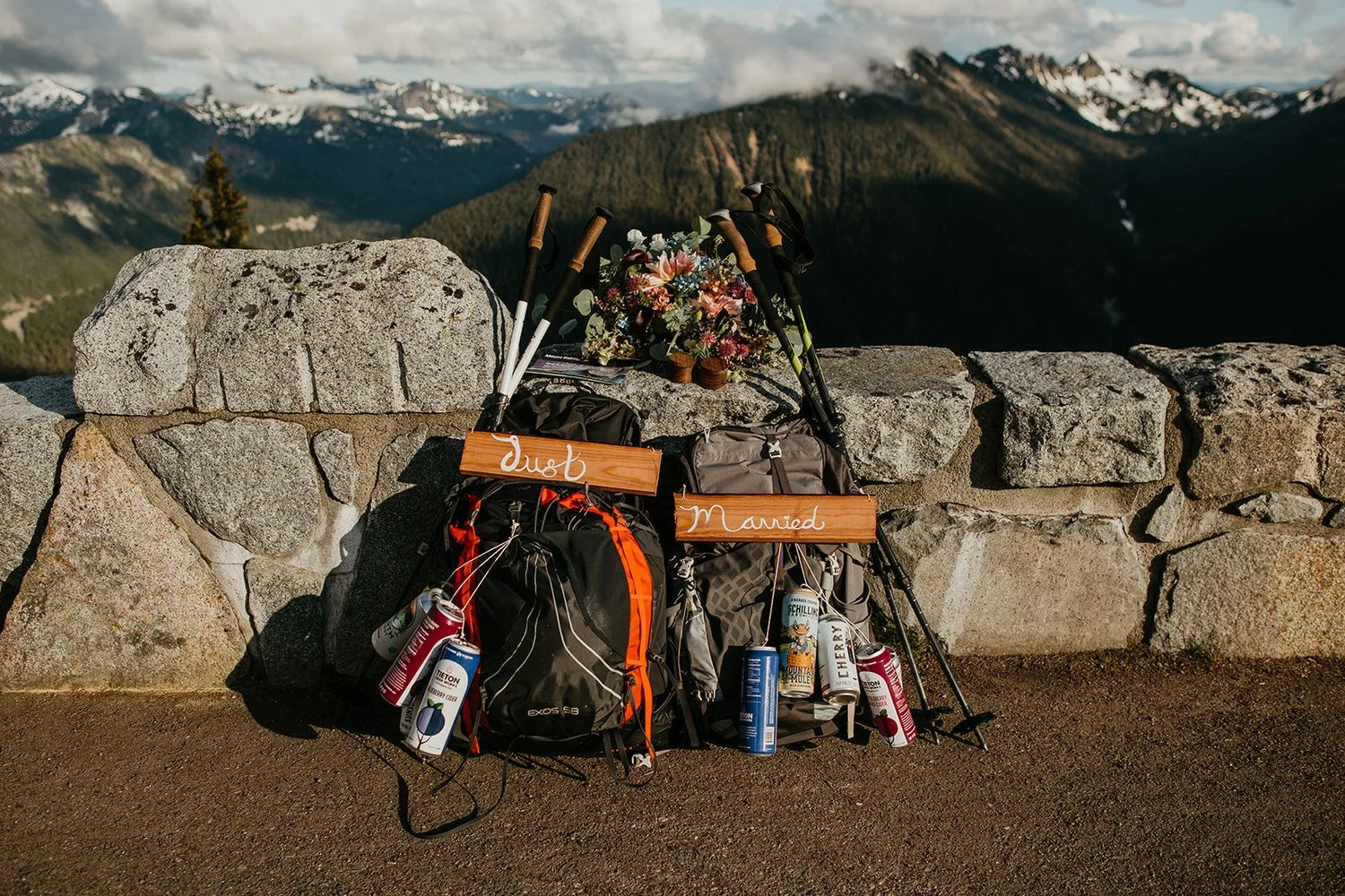 Two hiking backpacks with “just married” signs, trekking poles, and florals overlooking a mountain valley