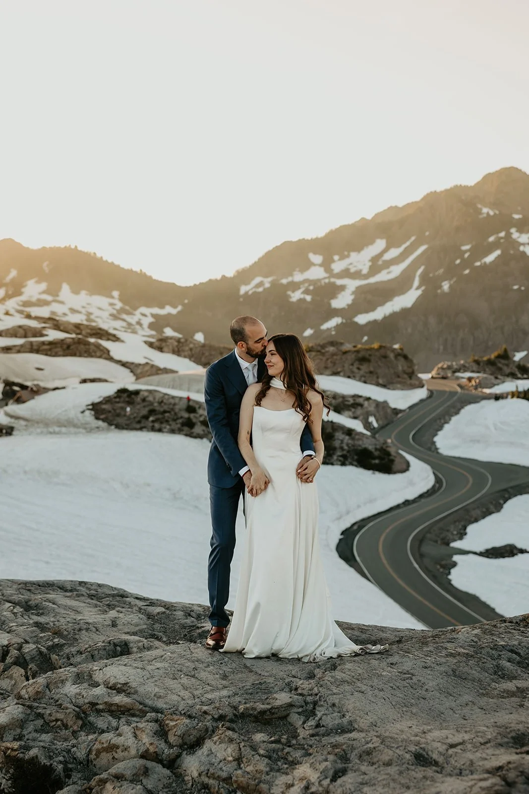 A couple embraces on a rocky overlook above a winding mountain road, surrounded by snow patches and rugged alpine terrain.