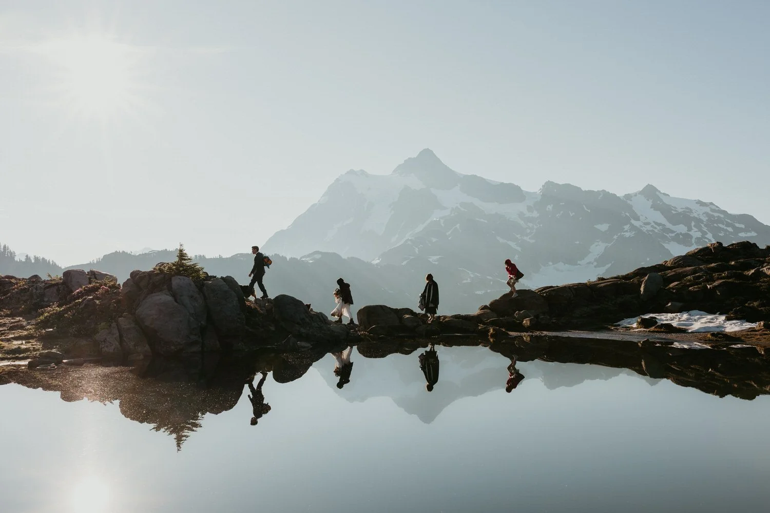 Guests walking along a rocky alpine trail beside a still lake reflecting Mount Shuksan at sunrise