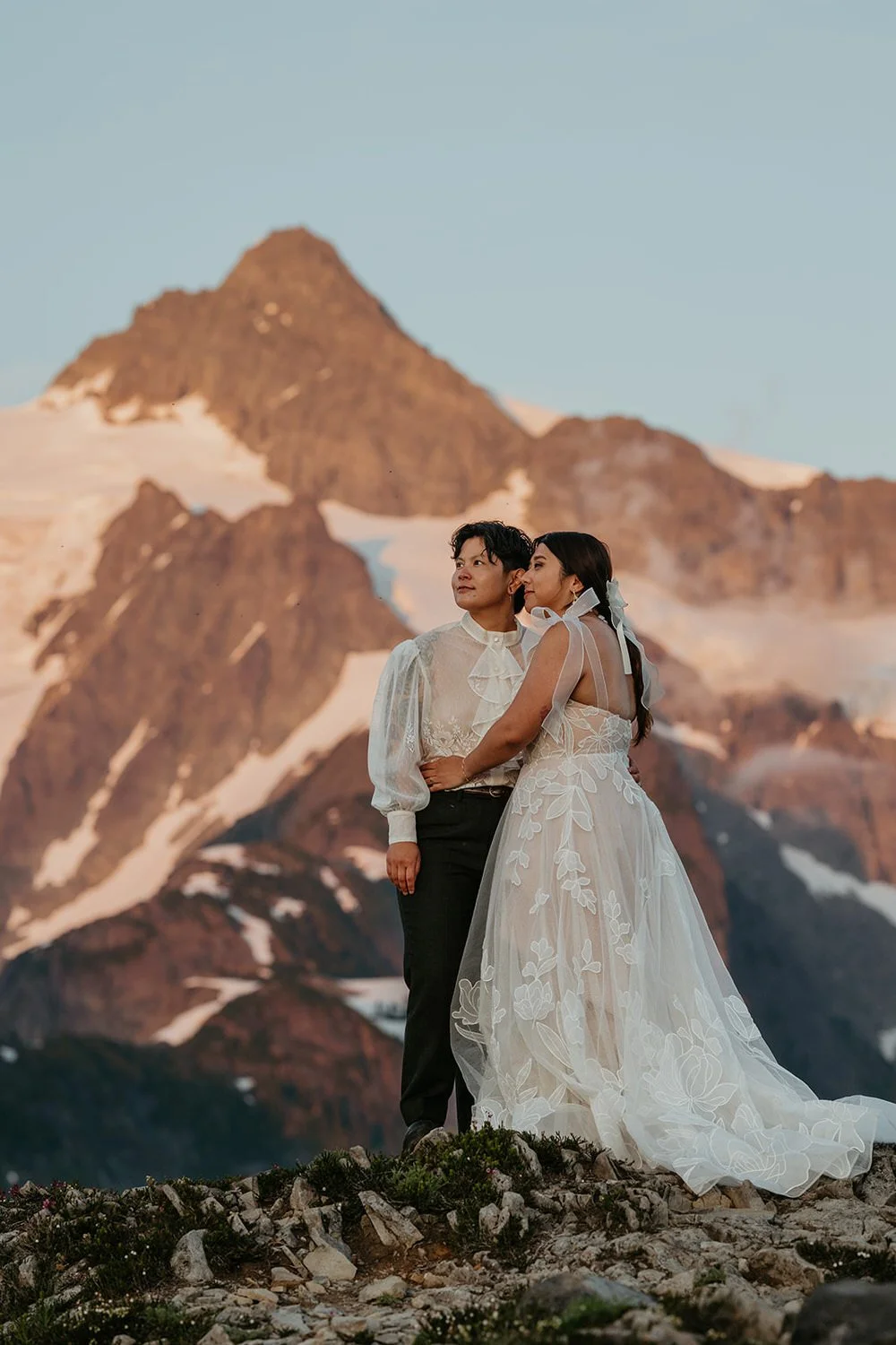 A queer couple stands together on a rocky alpine ridge at golden hour, with a rugged, snow-dusted mountain rising behind them.