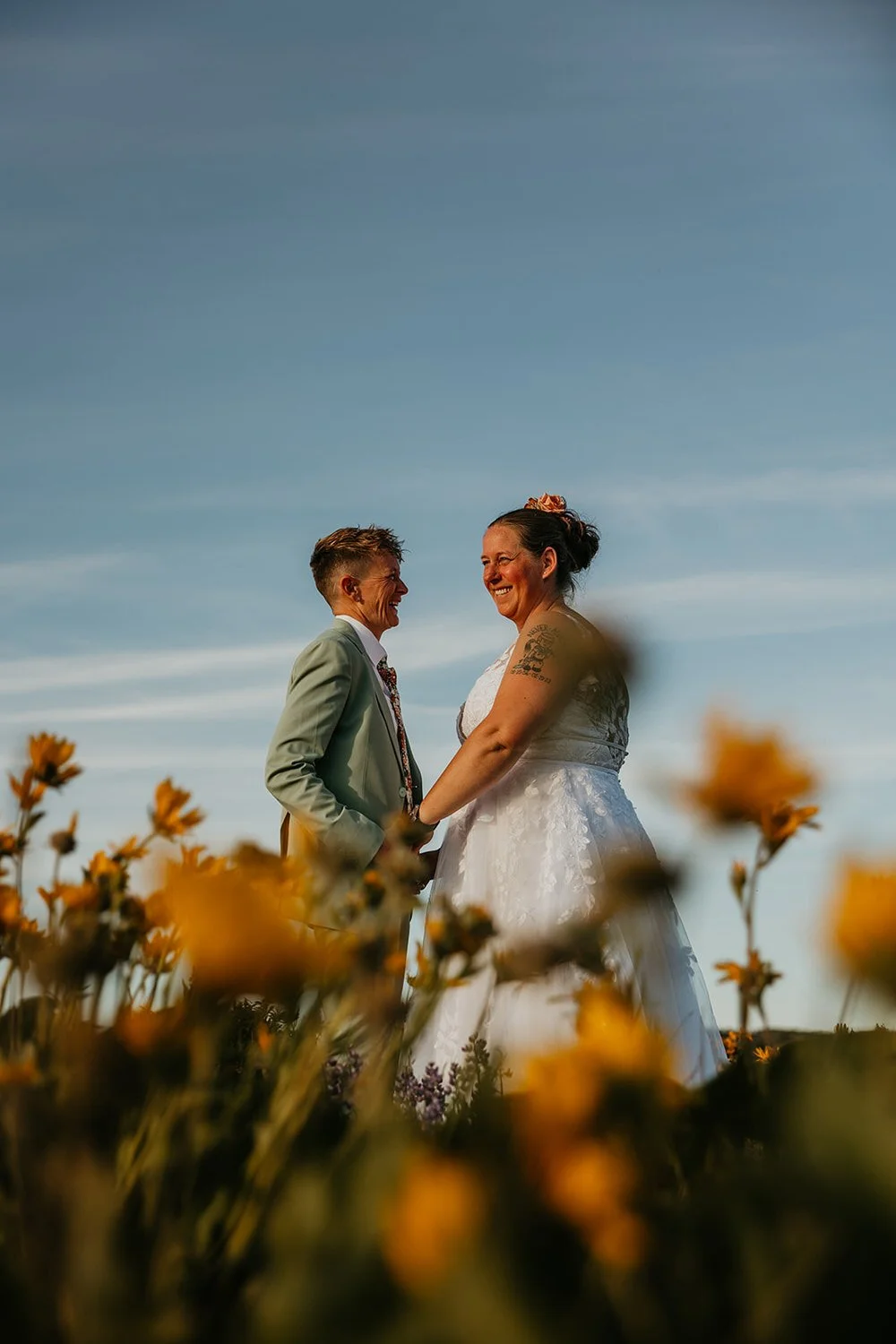 Two brides holding hands and laughing together in a field of golden wildflowers under a bright blue sky