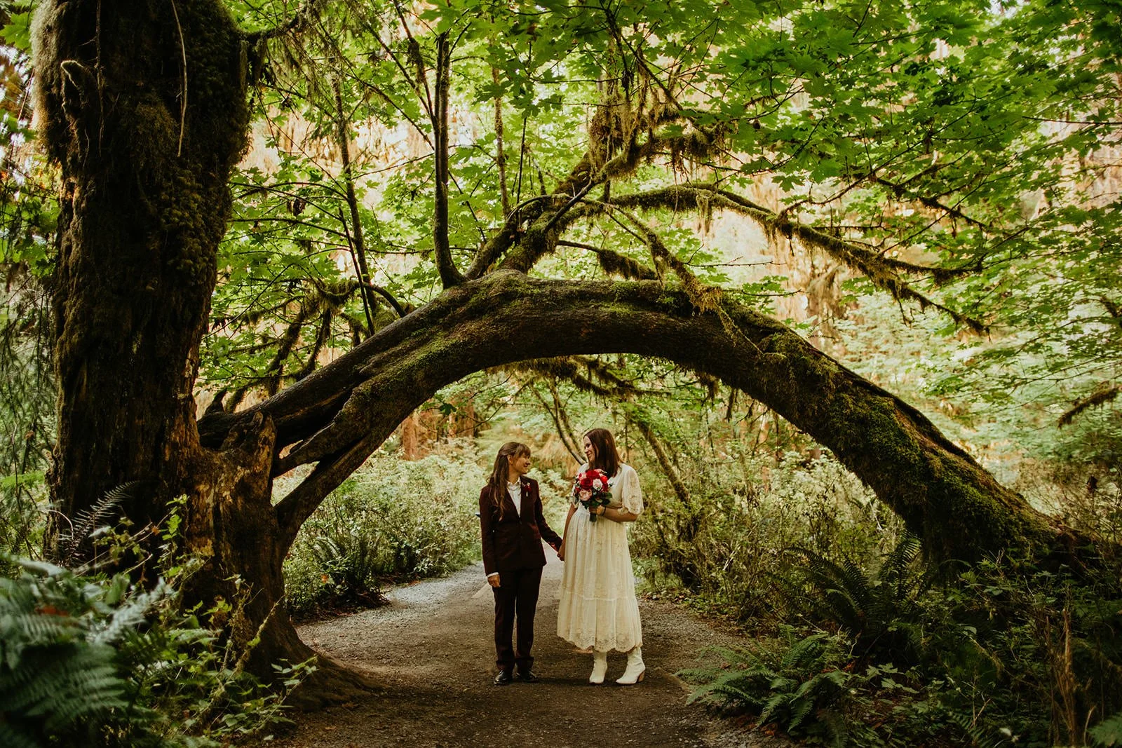 Two brides holding hands beneath a sweeping, curved tree along a quiet forest trail, surrounded by dense greenery and mossy branches.
