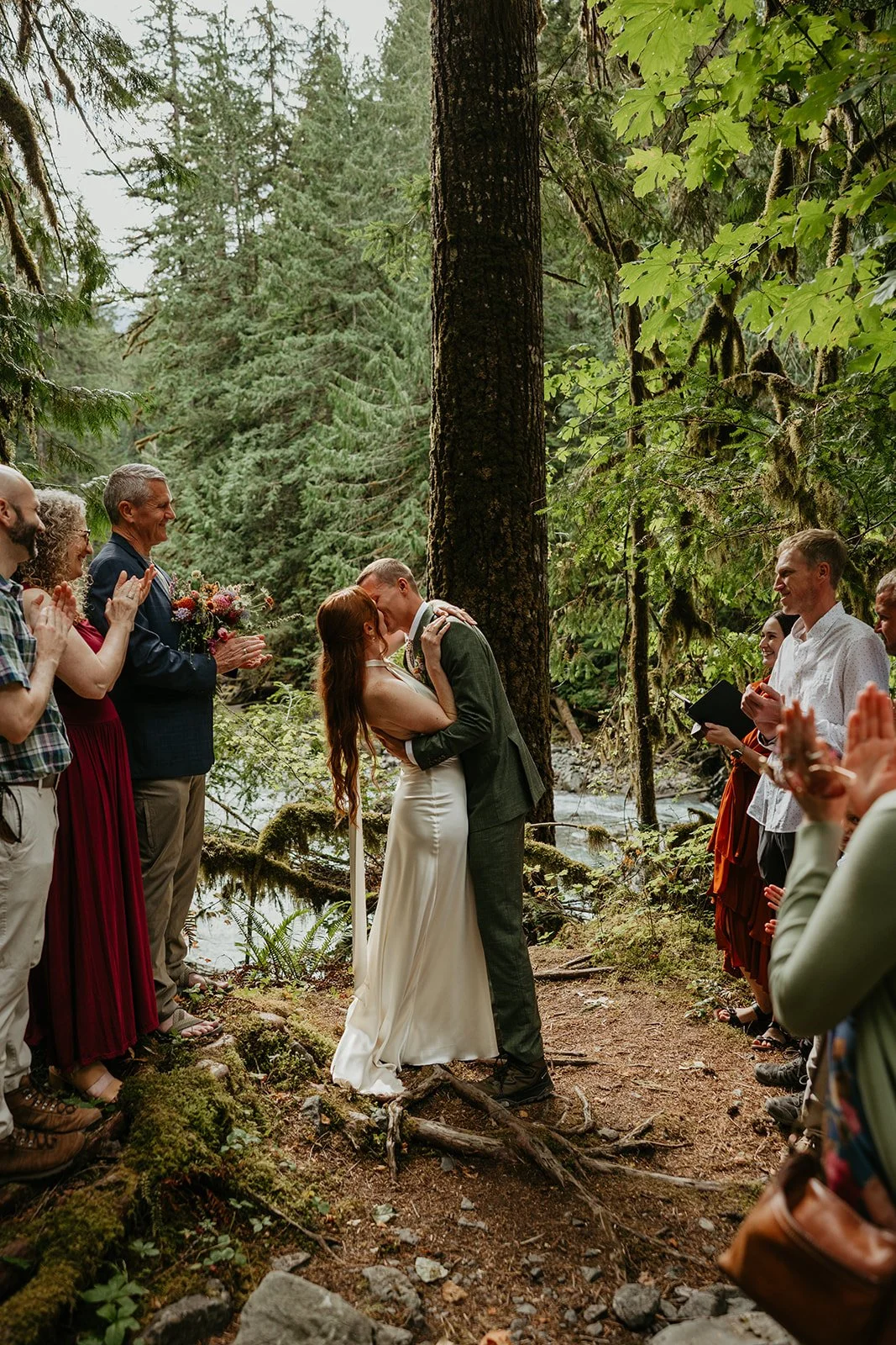 Couple exchanging vows beneath towering evergreen trees while friends and family watch during a forest elopement ceremony.