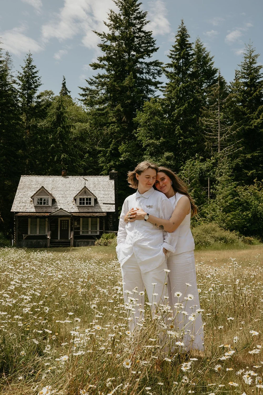 A queer couple standing together in a wildflower meadow, a small historic cabin and evergreen forest behind them.