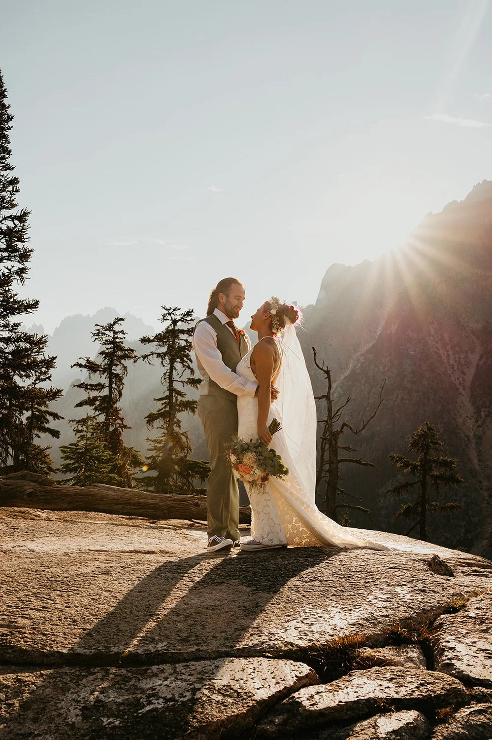 Bride and groom embrace while the sun shines on them from behind, bouquet in hand and scattered trees all around
