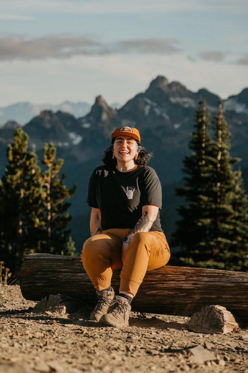 A woman sitting on a log outdoors, smiling, with mountains and trees in the background.