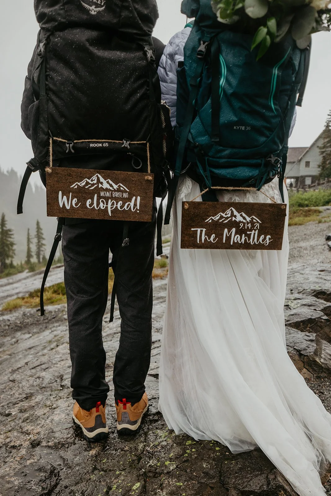 Back view of an eloping couple wearing backpacks during a Washington elopement, with wooden signs reading “We eloped” and “The Marvilles” hanging from their packs.