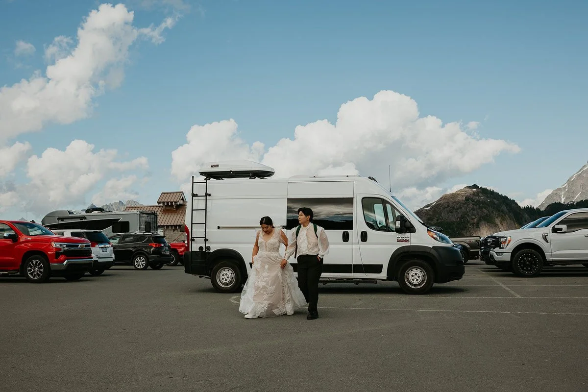 A bride and groom walking together in a parking lot, with various cars and an RV trailer in the background, and mountains in the distance.