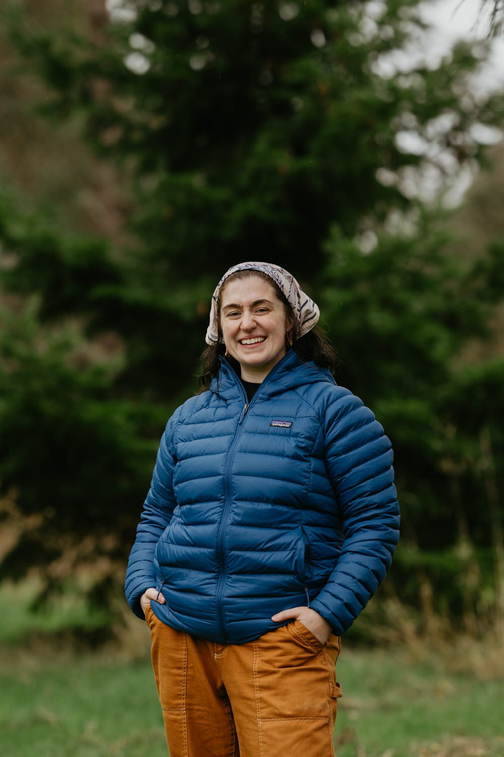 A woman standing outdoors in a green setting, smiling with hands in pockets, wearing a blue puffer jacket, tan pants, and a patterned headscarf.