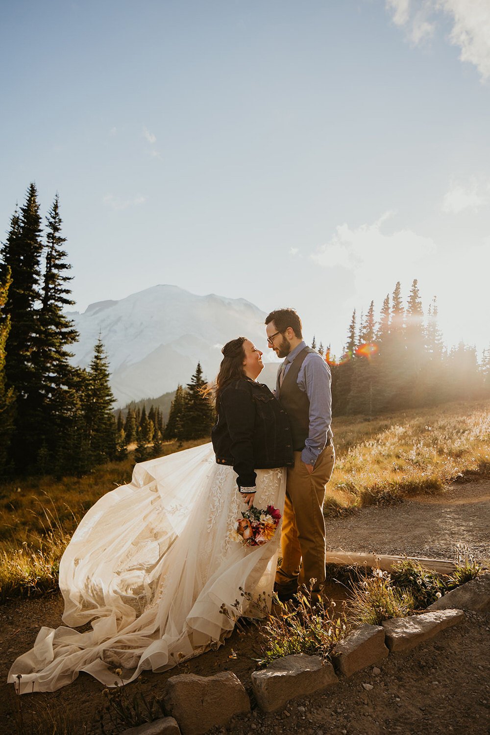 A couple stands close together at sunset in an alpine meadow, with Mount Rainier glowing softly behind them and tall evergreen trees nearby.