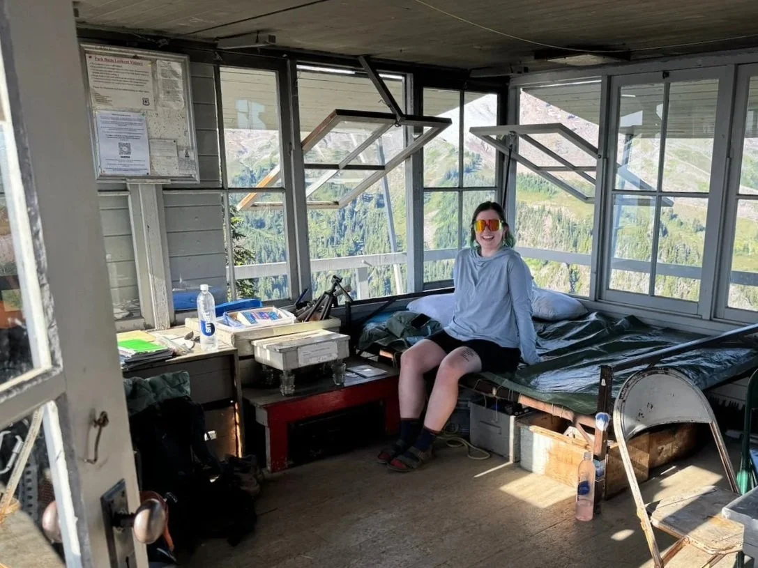 A woman with short dark hair wearing a light gray hoodie, black shorts, socks, sandals, and orange sunglasses, sitting on a bed inside a small room with large windows showing a mountainous landscape.