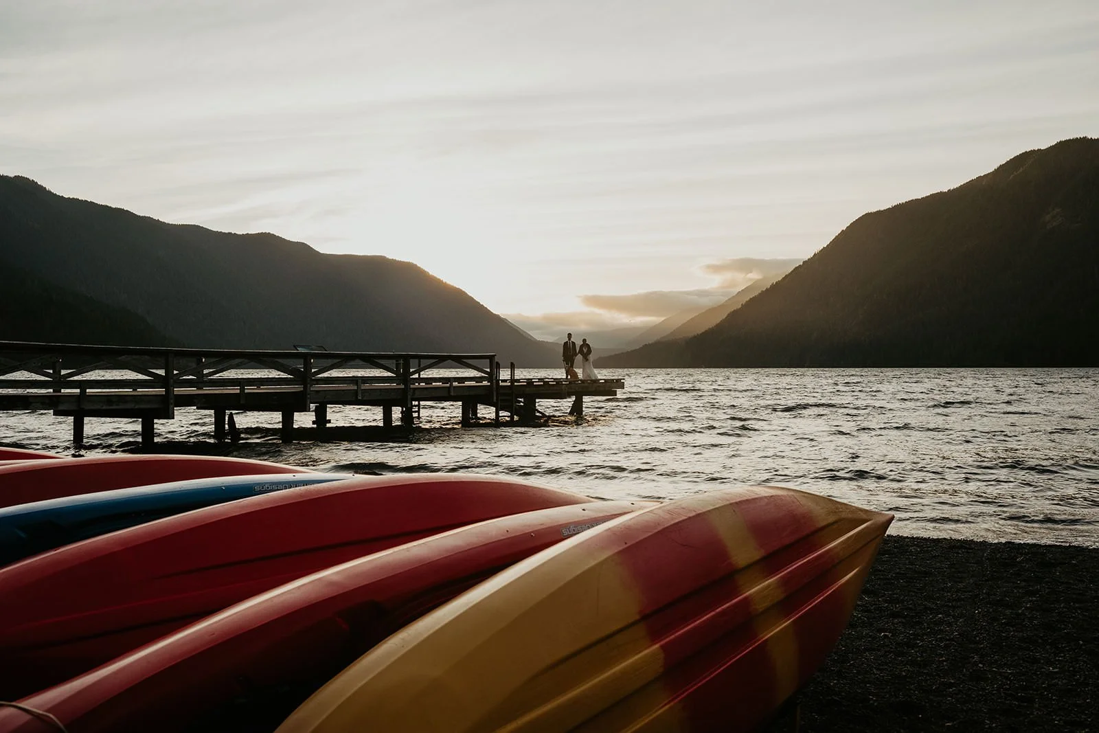 Couple standing at the end of a wooden dock at sunset, mountain silhouettes surrounding a Washington lake with colorful kayaks in the foreground.