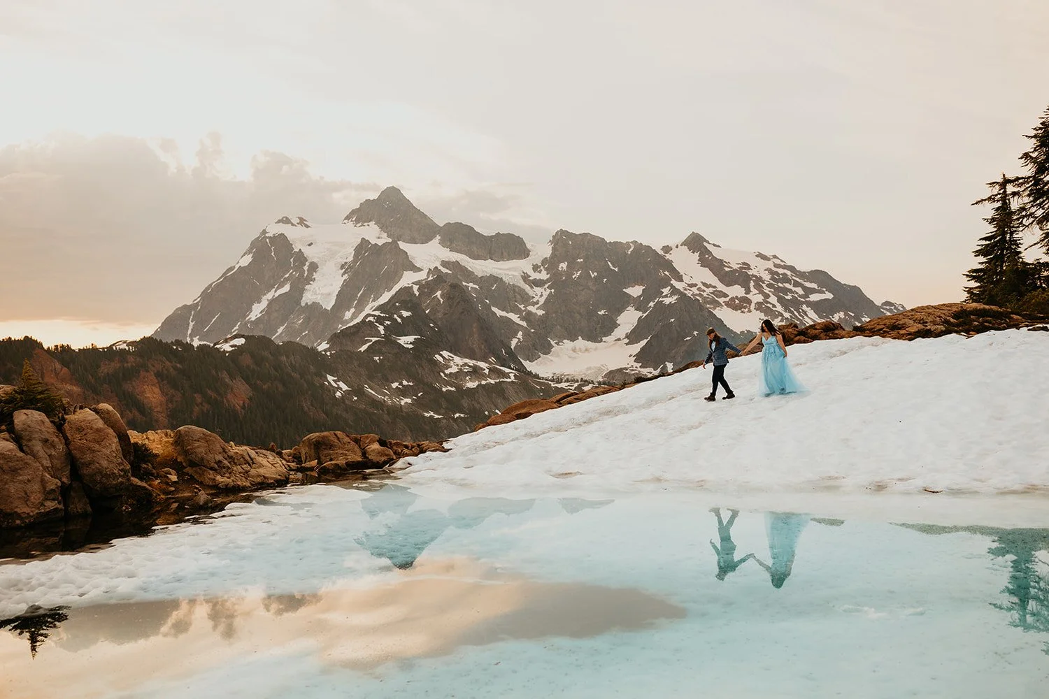 Eloping couple walking hand in hand across a snowy alpine basin with jagged, glacier covered mountains reflected in an icy blue meltwater pool