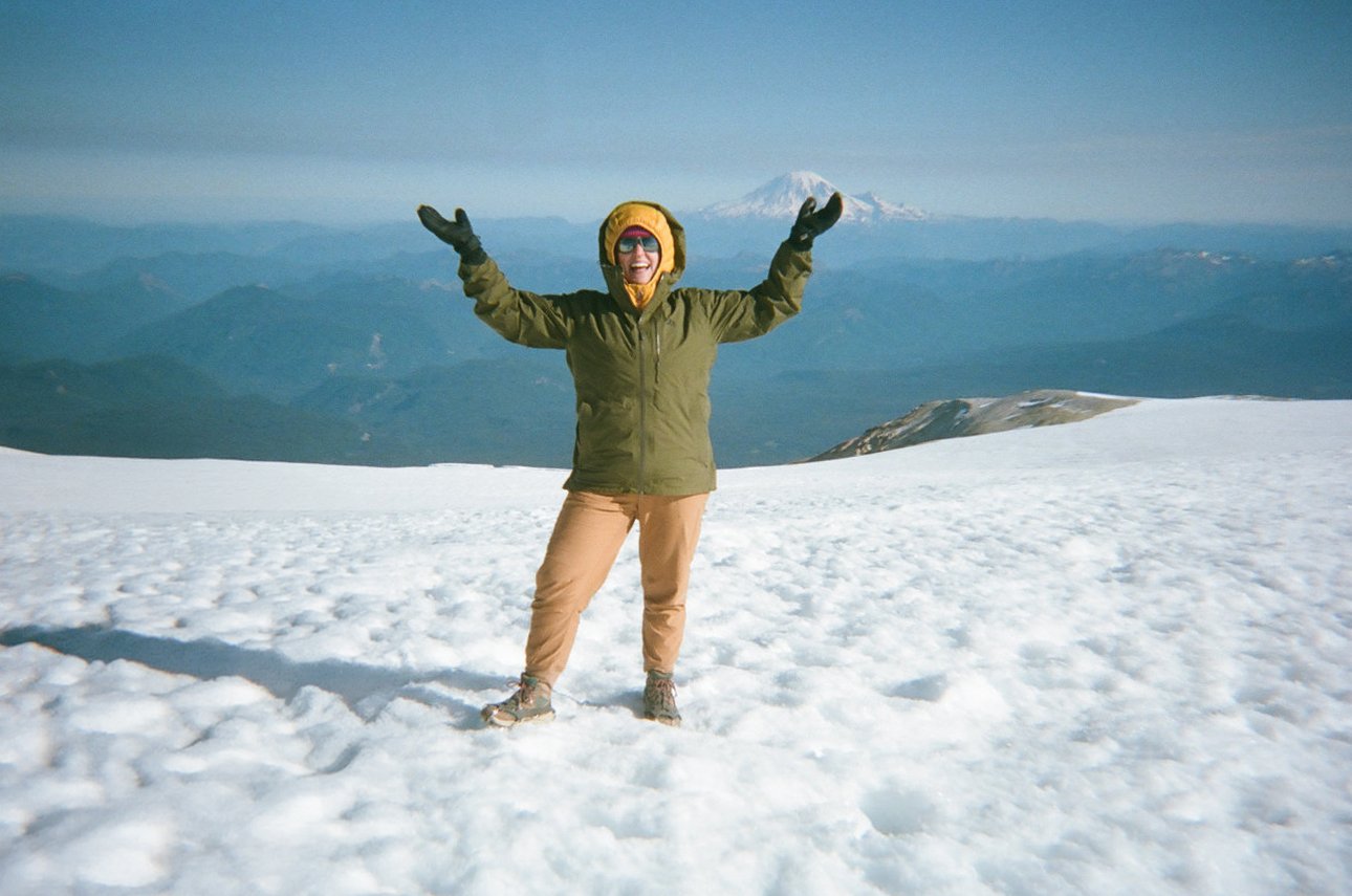 Person standing on snow-covered mountain peak, raising arms with a view of distant mountains and a snow-capped volcano in the background.