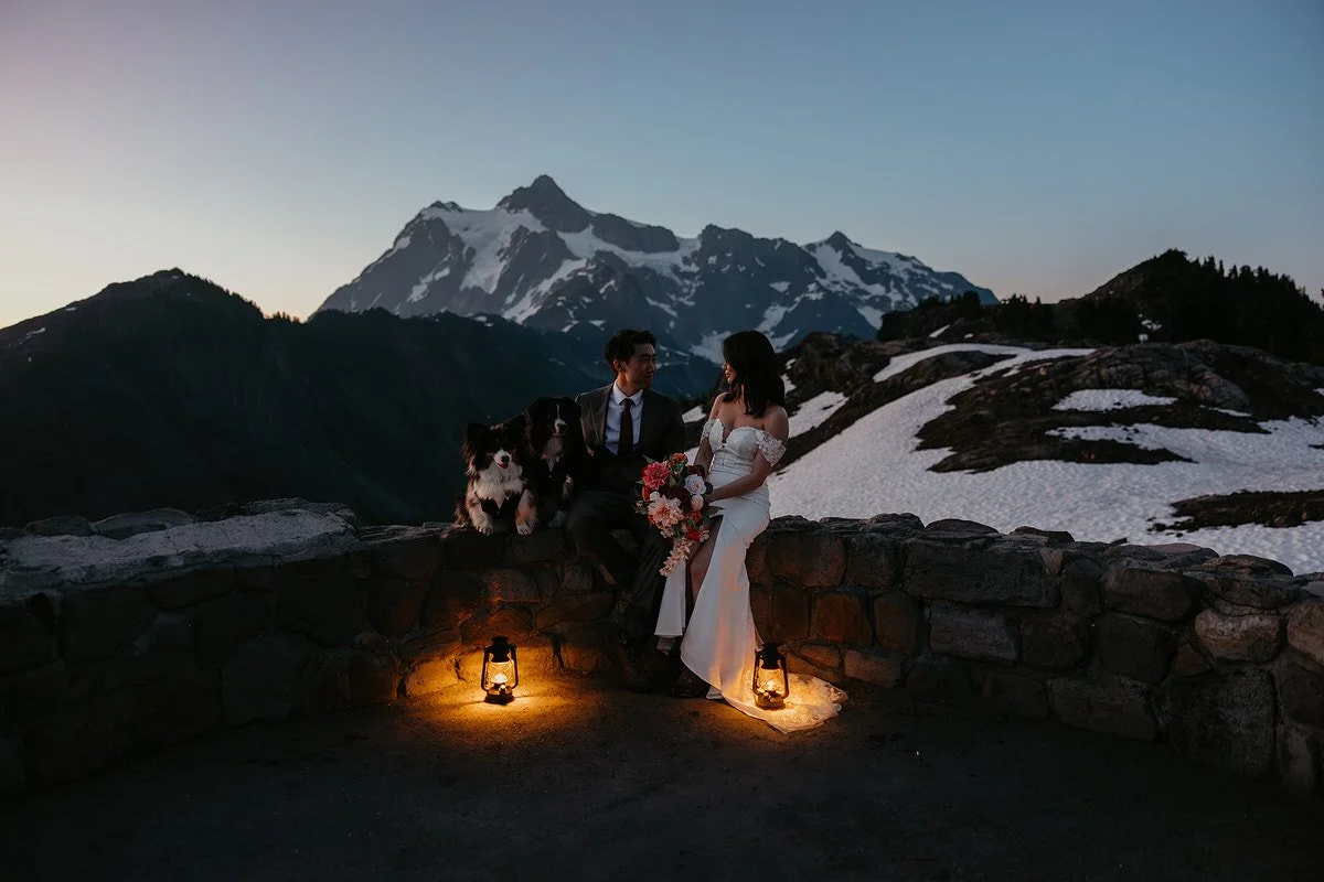 Couple sitting on a stone overlook at dusk with their dogs and lanterns, snowy mountain peak in the distance