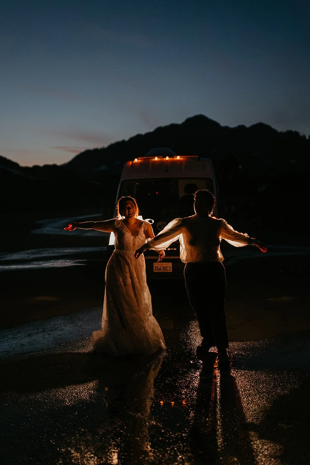 Eloping couple dancing barefoot in the rain at night in front of a camper van with headlights glowing on a mountain road.