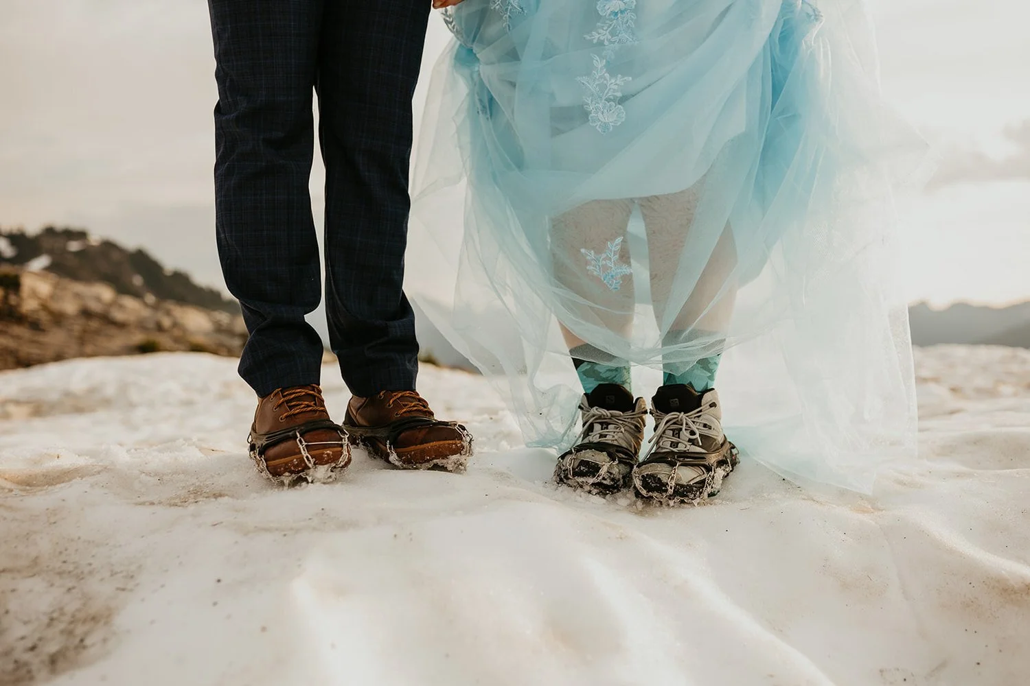 Close up of an eloping couple’s feet in the snow, showing hiking boots and crampons beneath a flowing blue wedding dress