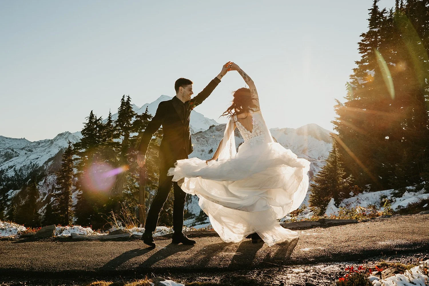 Couple twirling in a flowing wedding dress on a snowy Washington mountaintop, bright sun flare filtering through evergreen trees during a winter elopement.