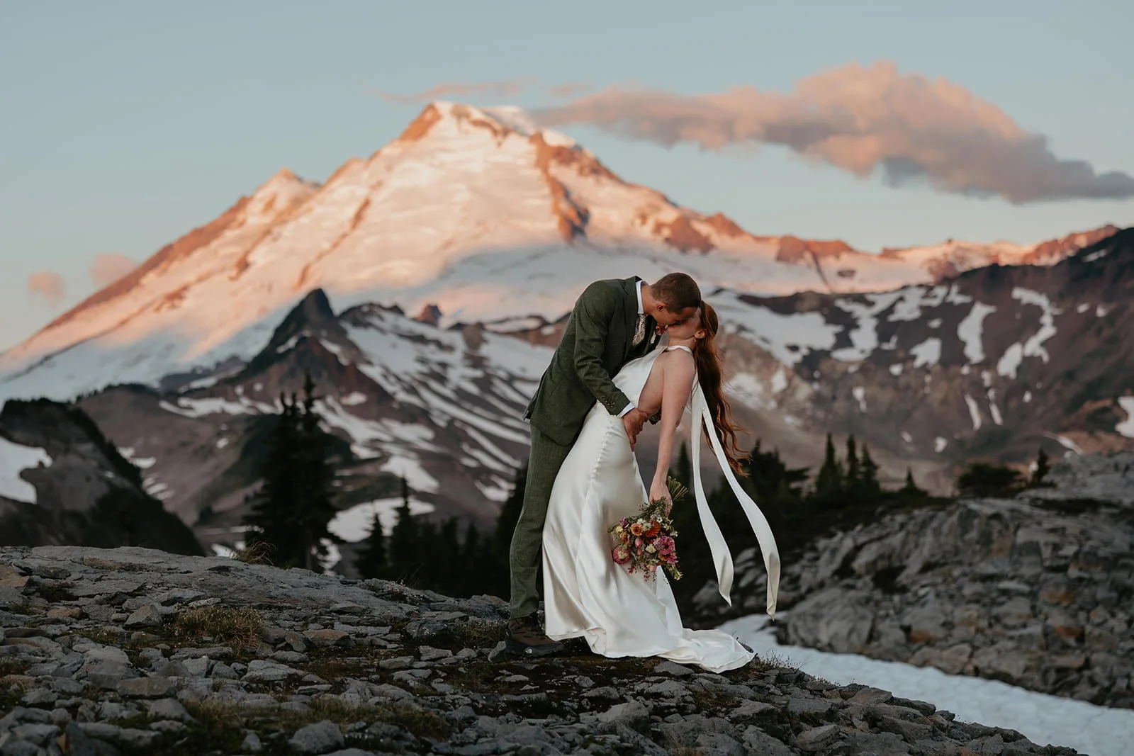 Couple embracing on rocky alpine terrain with Mount Rainier glowing in the background at sunset.