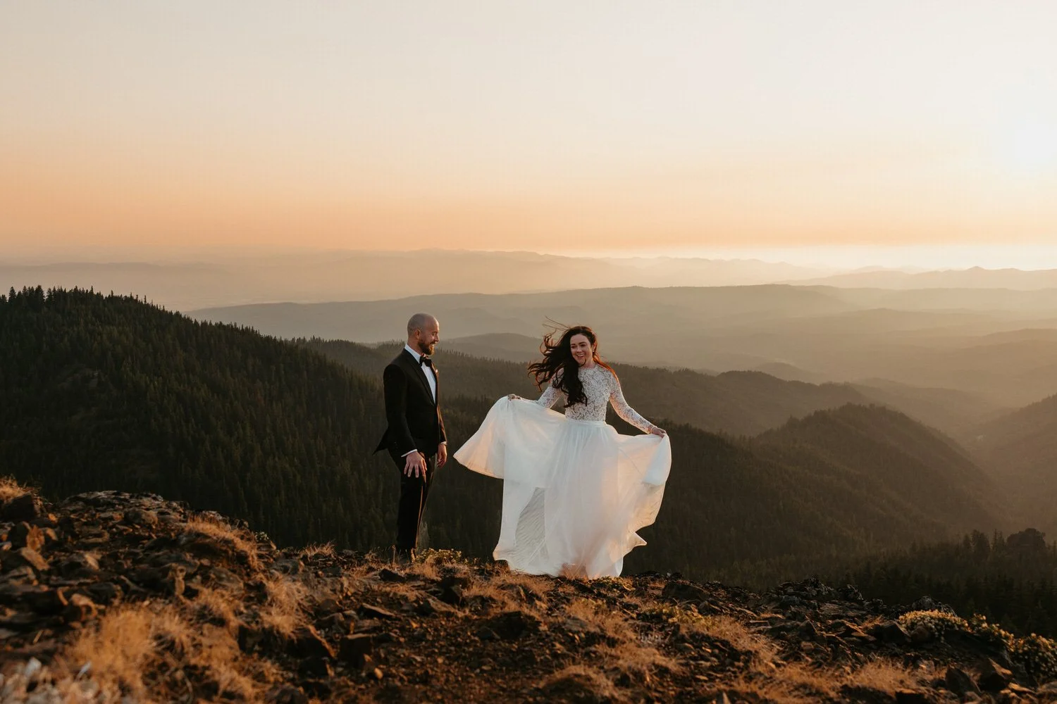 Bride twirling her dress on a mountaintop at golden hour while her partner watches, with layered mountain ridges fading into the distance.