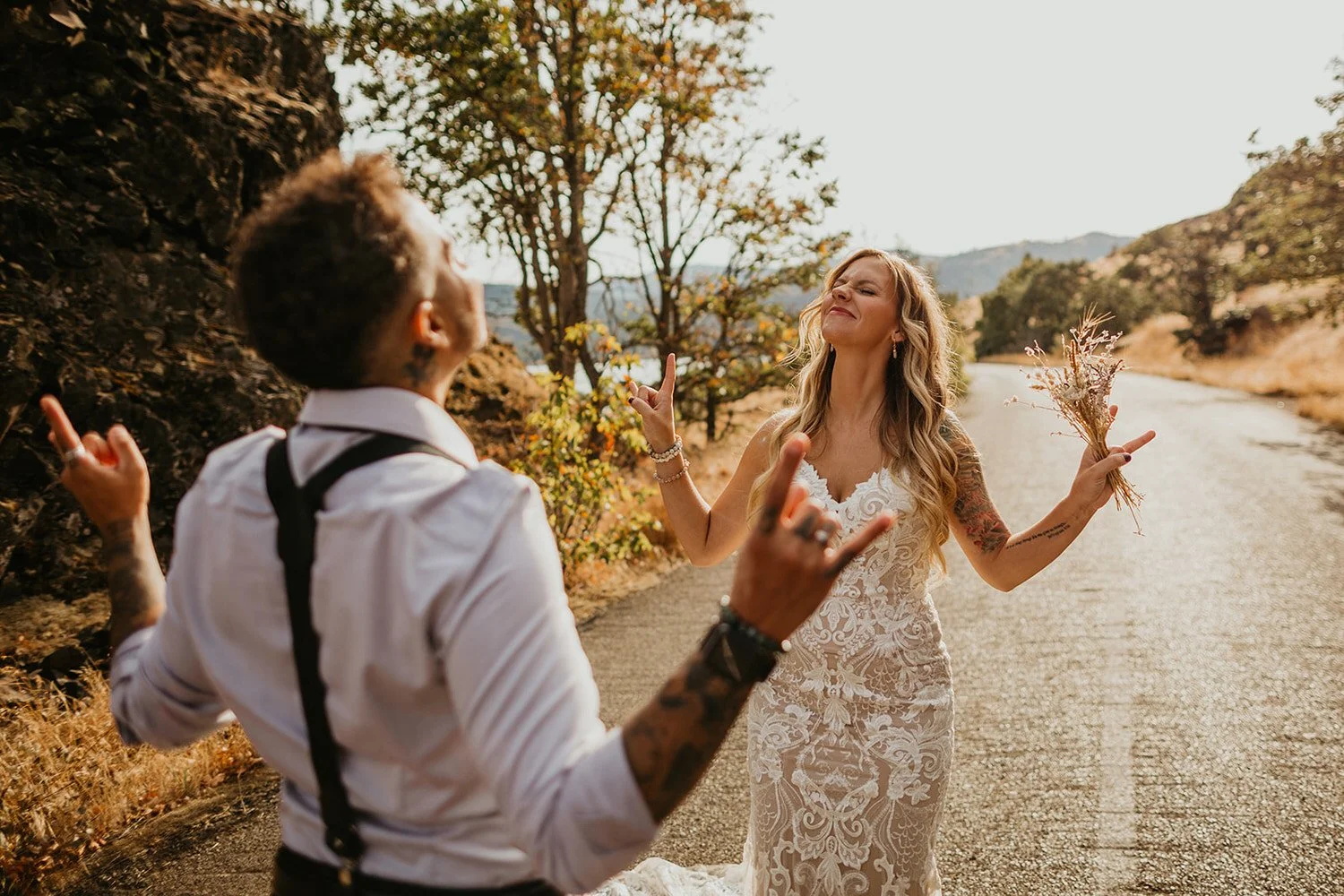 A couple dancing outdoors on a rural road during sunset, with the woman holding a bouquet of dried flowers and smiling, the man in a white shirt with suspenders, and trees with fall foliage in the background.