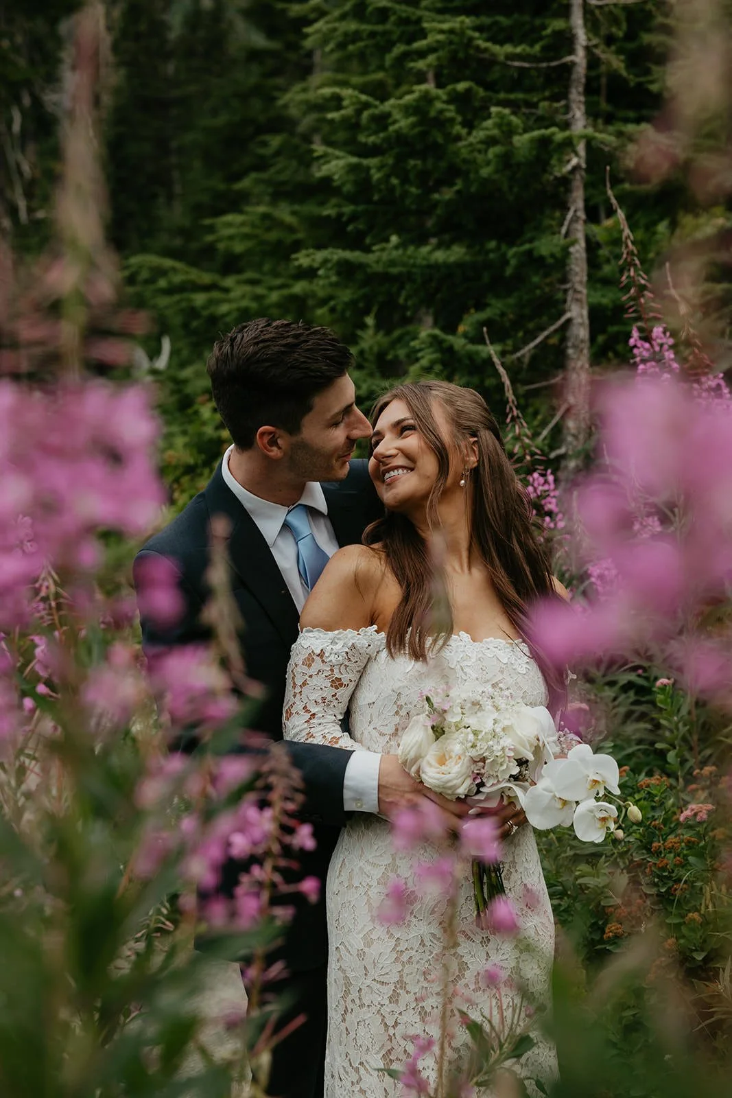 A couple embraces among tall pink wildflowers in a forest clearing, surrounded by lush green trees.
