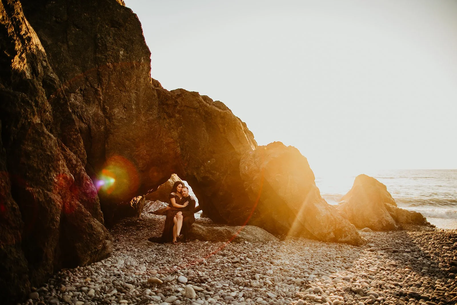Couple sitting beneath a natural rock arch on a rocky beach as sunlight flares through the opening near the ocean shoreline.