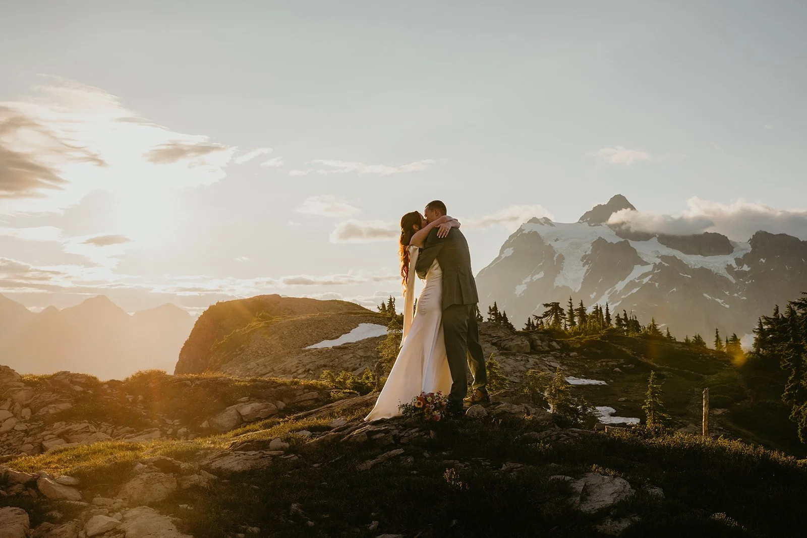 Couple embracing on a grassy alpine ridge at sunset with warm golden light spilling across Mount Baker in the background during their Washington elopement.