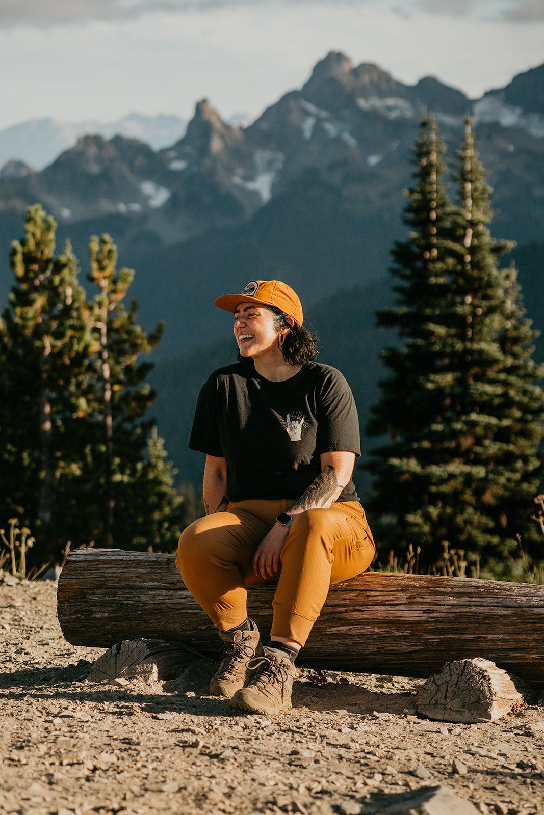 A woman sitting on a log in a mountainous forest area, smiling and enjoying the outdoors during daytime.