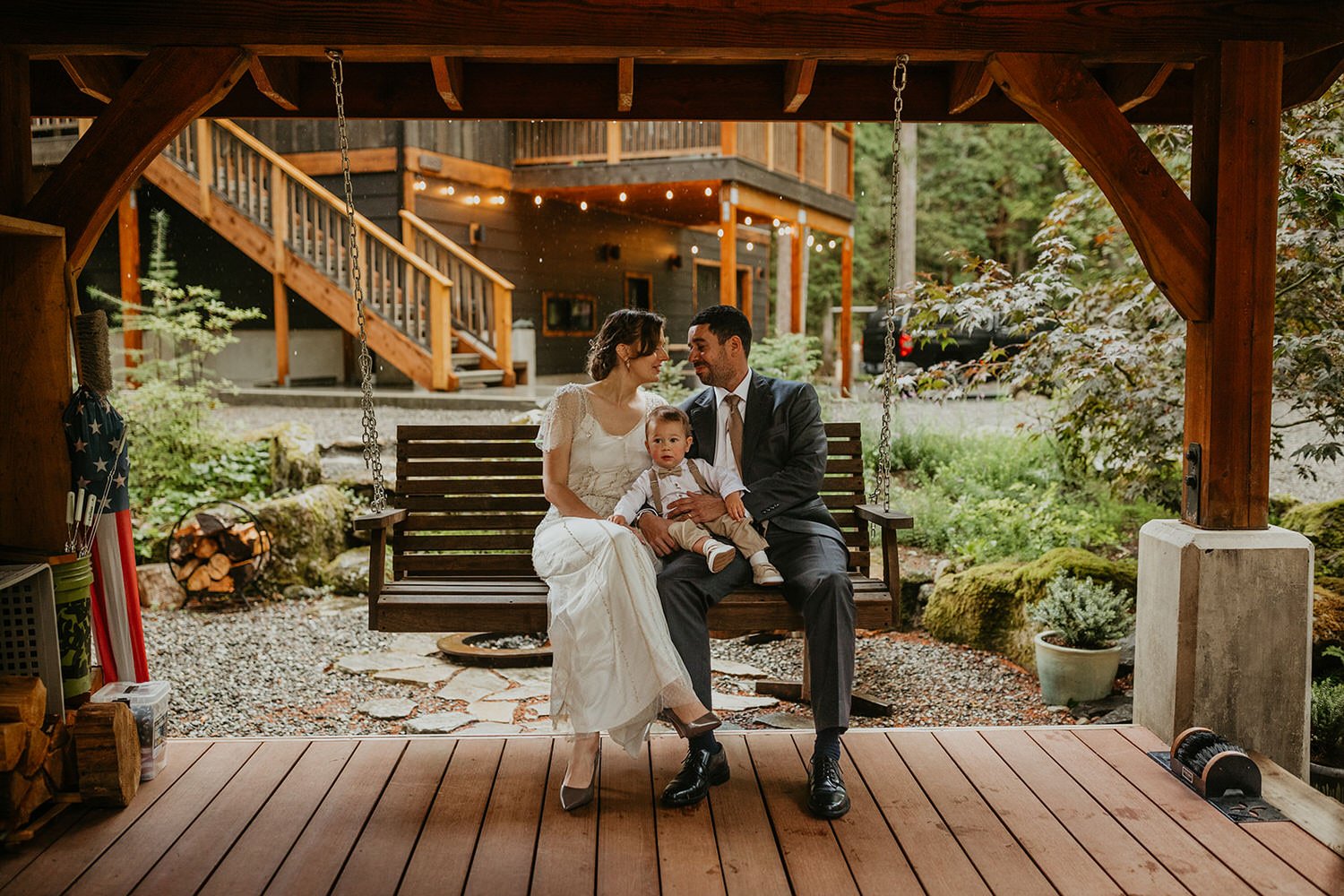 Couple in wedding attire sitting on a wooden porch swing with their baby during a Washington Airbnb elopement, string lights and a multi-level cabin visible in the background