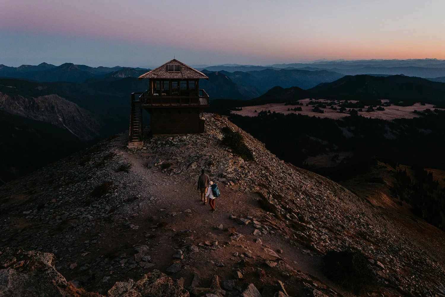 Couple hiking toward a fire lookout at dusk with expansive mountain views and layered ridgelines at sunset