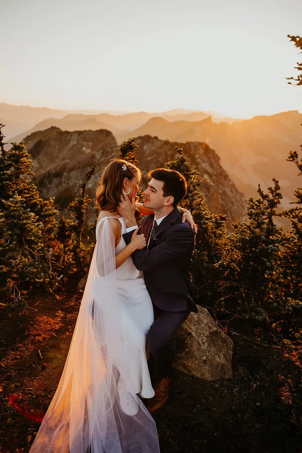 Sunset glows behind an elopement couple in the North Cascades as the bride sits on the grooms lap and they share a moment together