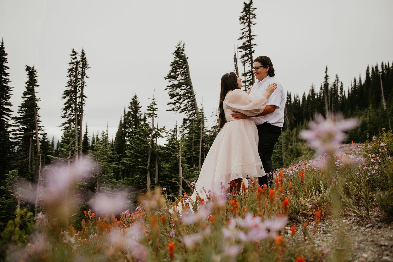 Couple embracing in alpine meadow surrounded by pink and orange wildflowers; coniferous forest and dramatic peaks behind
