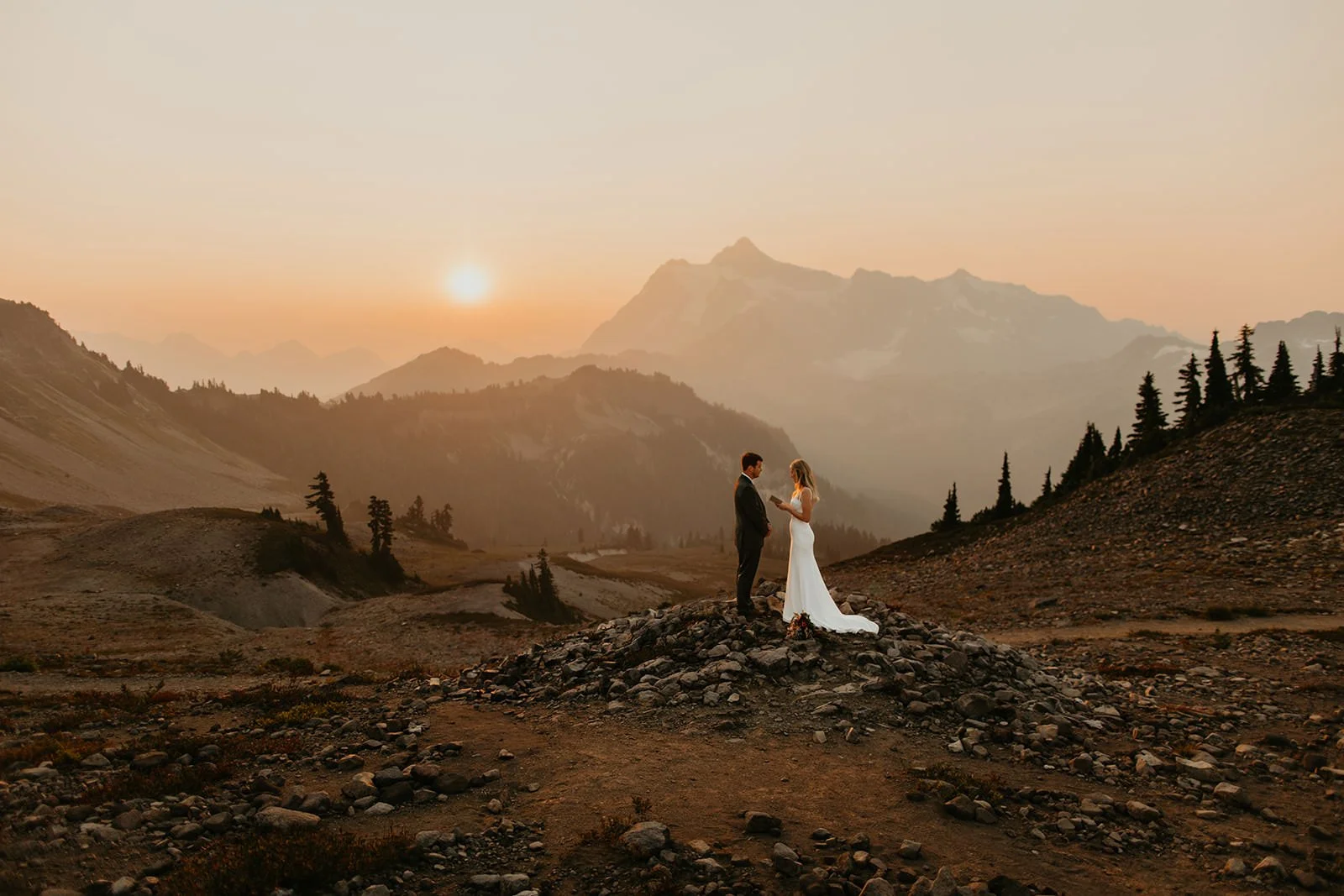 Couple standing on a rocky rise at sunset, surrounded by rolling alpine terrain and distant mountain peaks.