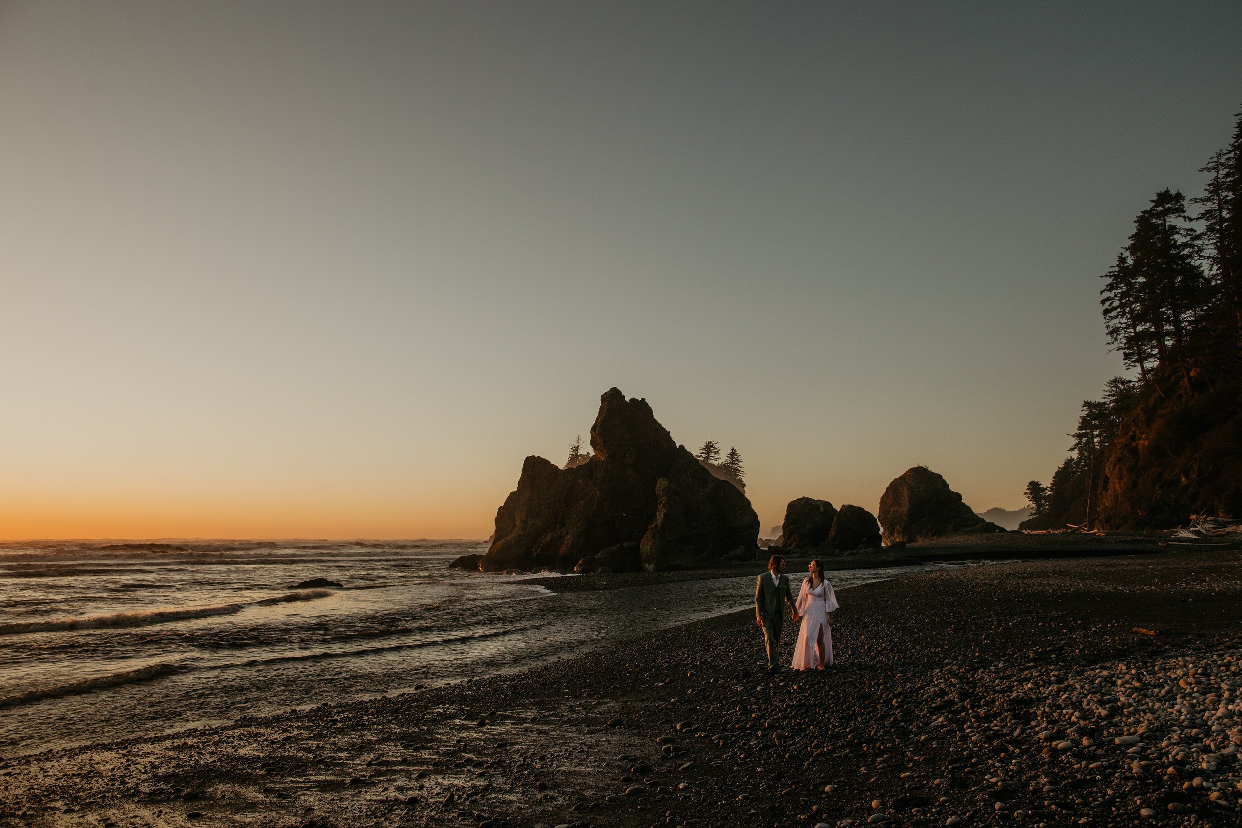 Couple walking along a rocky shoreline at sunset, dark sea stacks silhouetted against the sky as waves break along the shore.