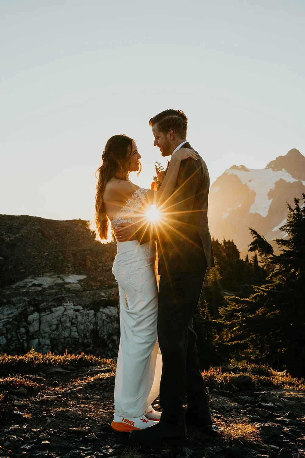 A couple stands close during a sunset mountain elopement, with warm sunlight flaring between them and jagged alpine peaks glowing in the background.