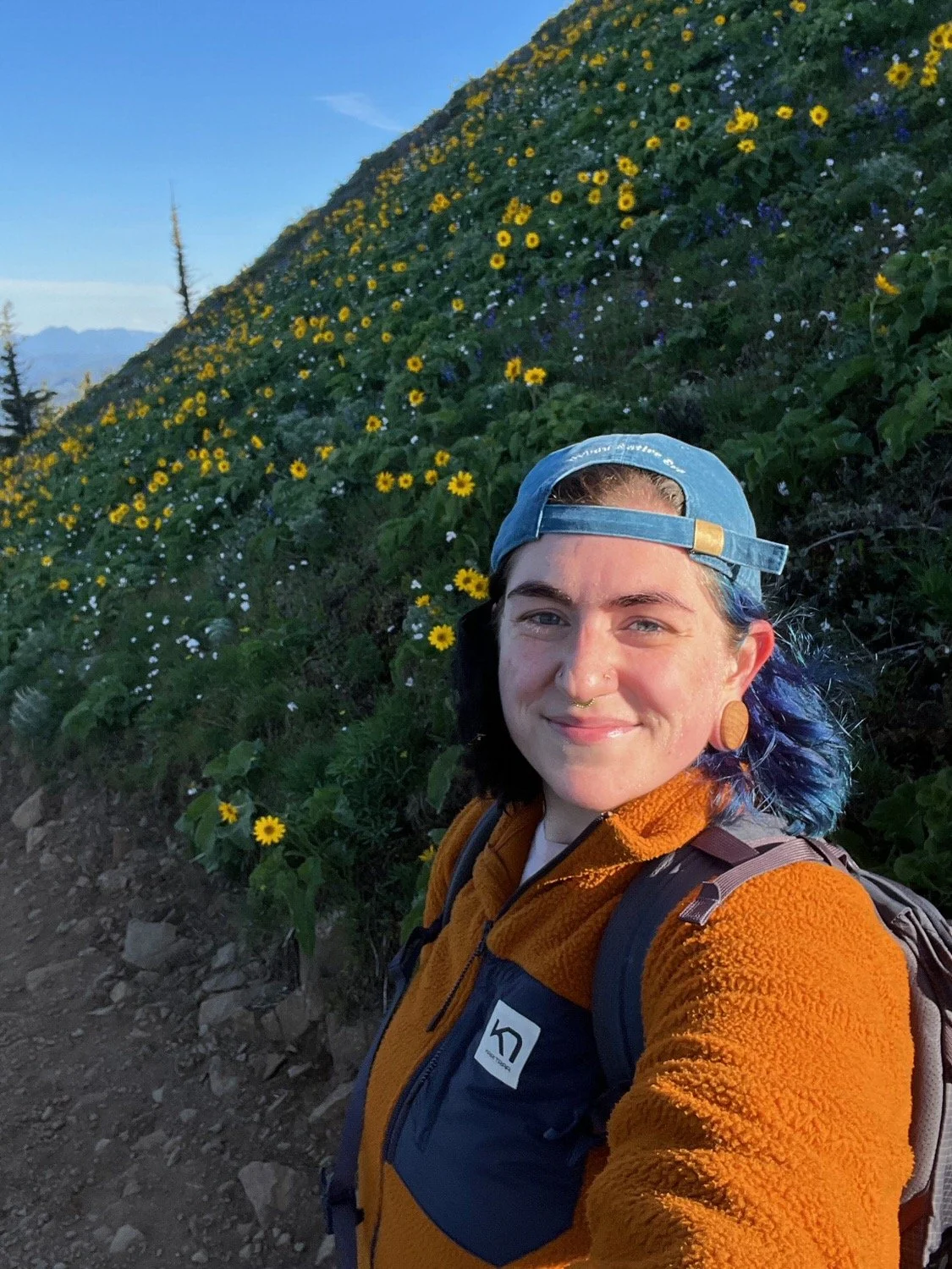 Van smiling at camera wearing blue cap and orange fleece jacket with backpack; standing on hillside densely covered in yellow and white wildflowers with coniferous forest on mountain slopes behind