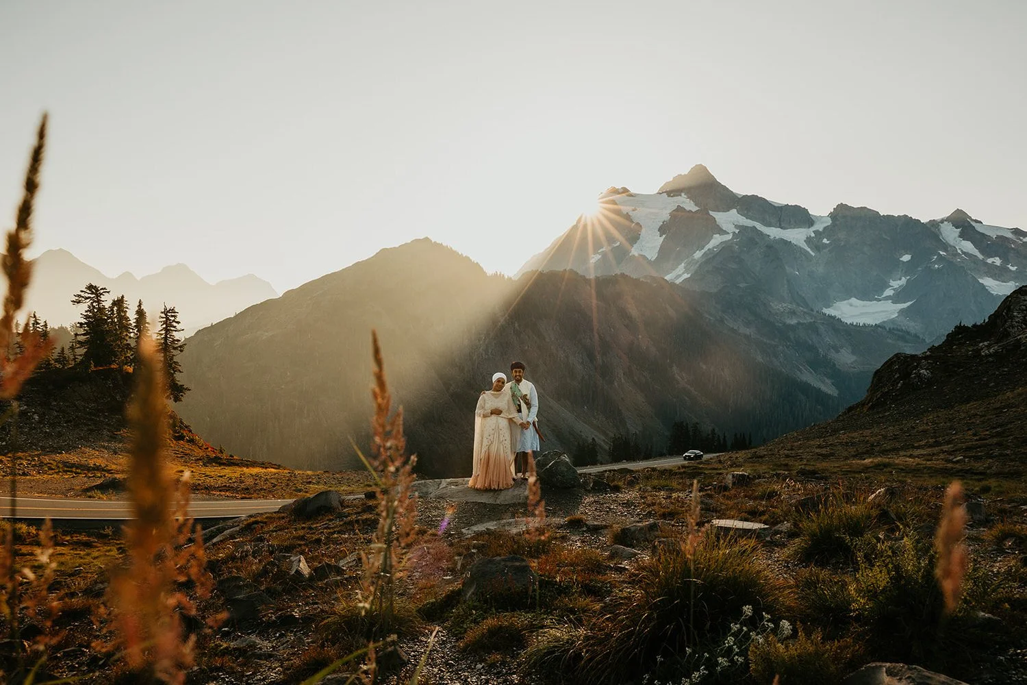 Eloping couple standing beside a mountain road at sunrise with golden light streaming over alpine peaks and wild grasses in the foreground