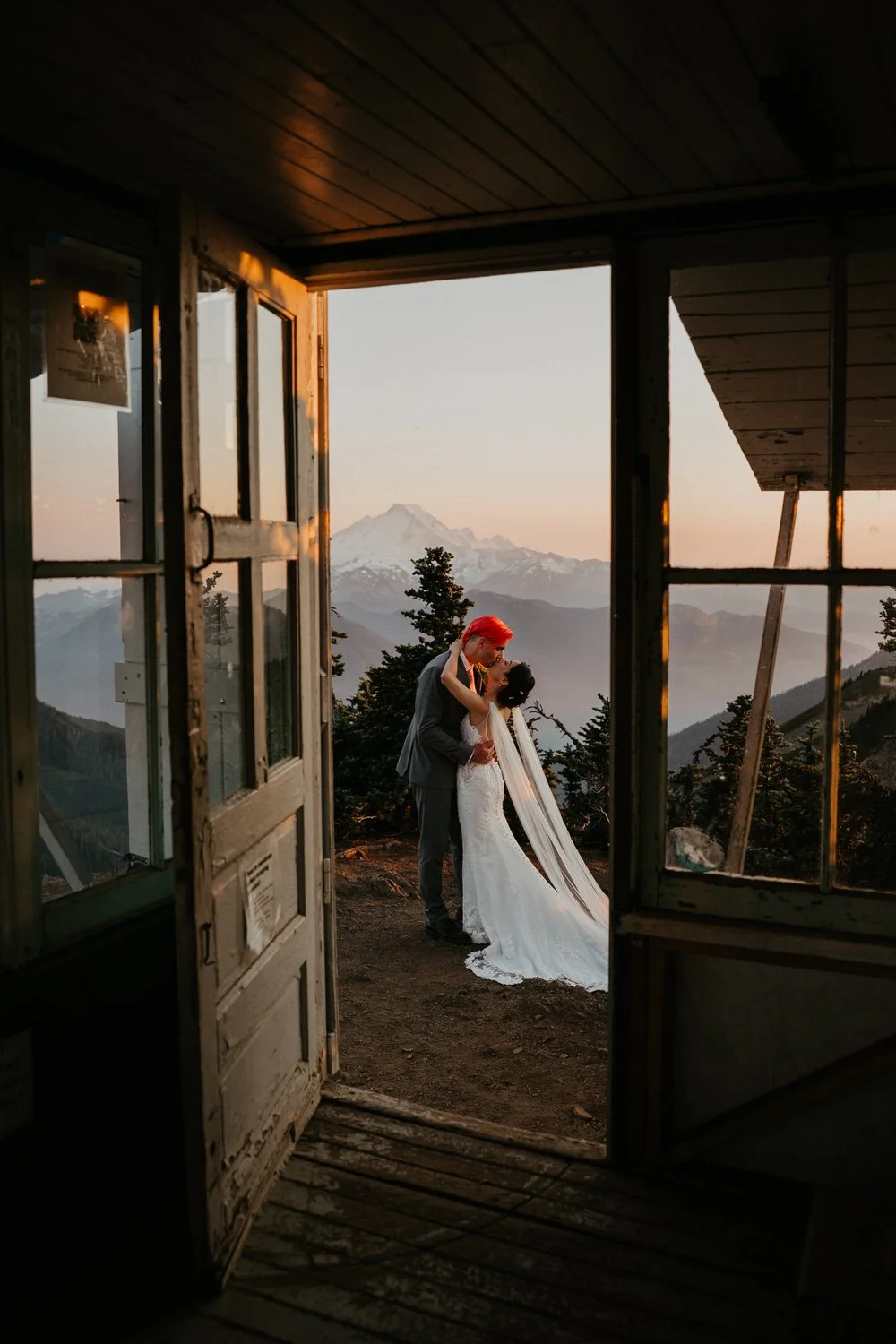 A couple kisses just outside a rustic mountain lookout structure, framed by open doors with distant peaks and soft evening light beyond.