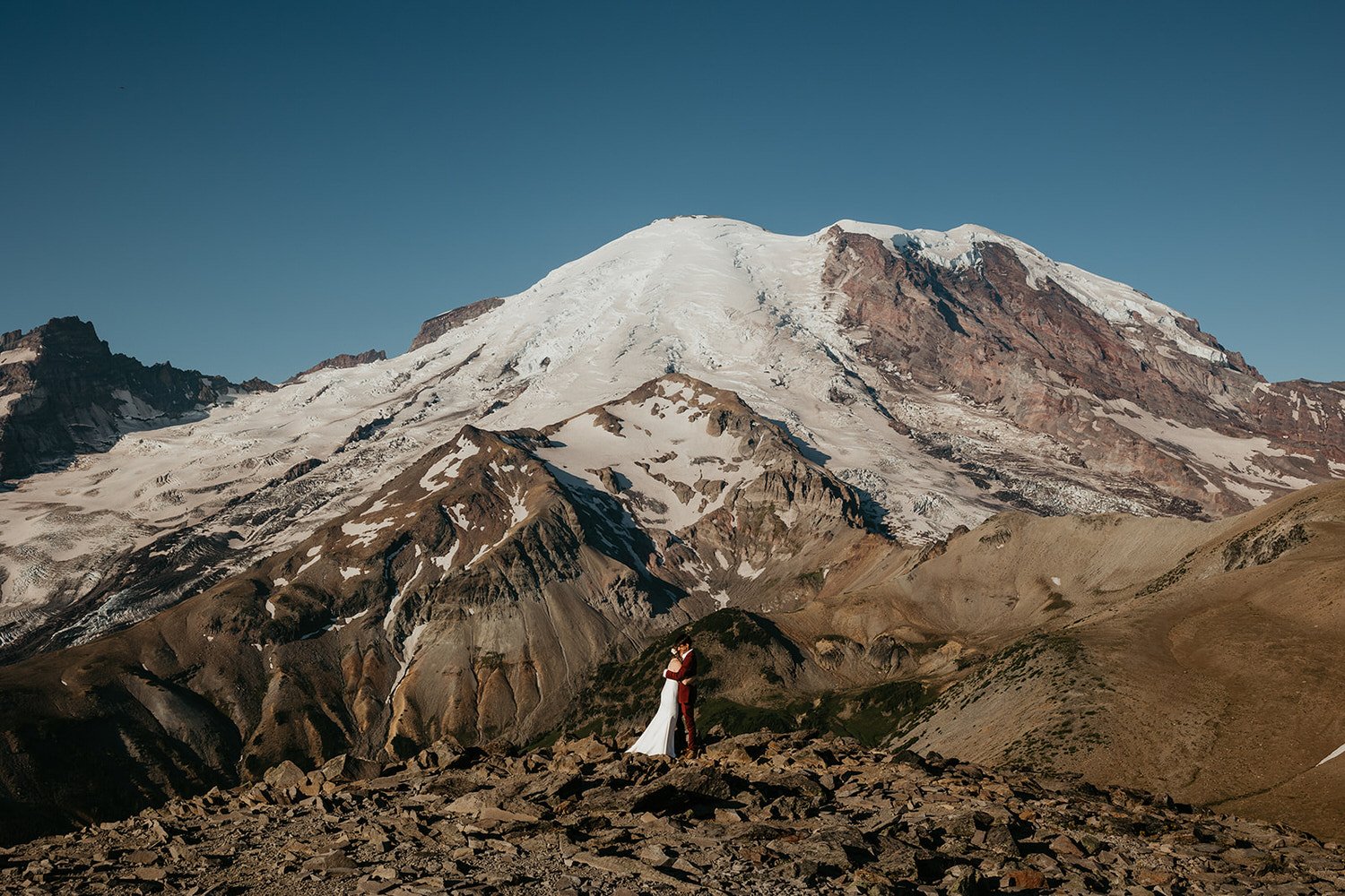 Eloping couple standing on a rocky overlook with sweeping alpine terrain and a glacier-covered peak behind them