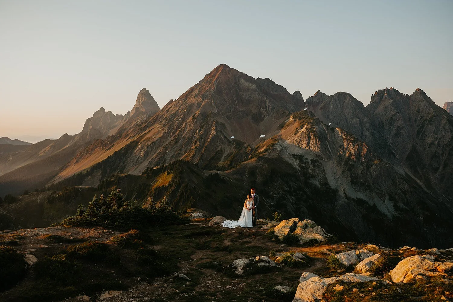 Eloping couple standing on an alpine ridge surrounded by jagged mountain peaks during golden hour with soft light on the landscape