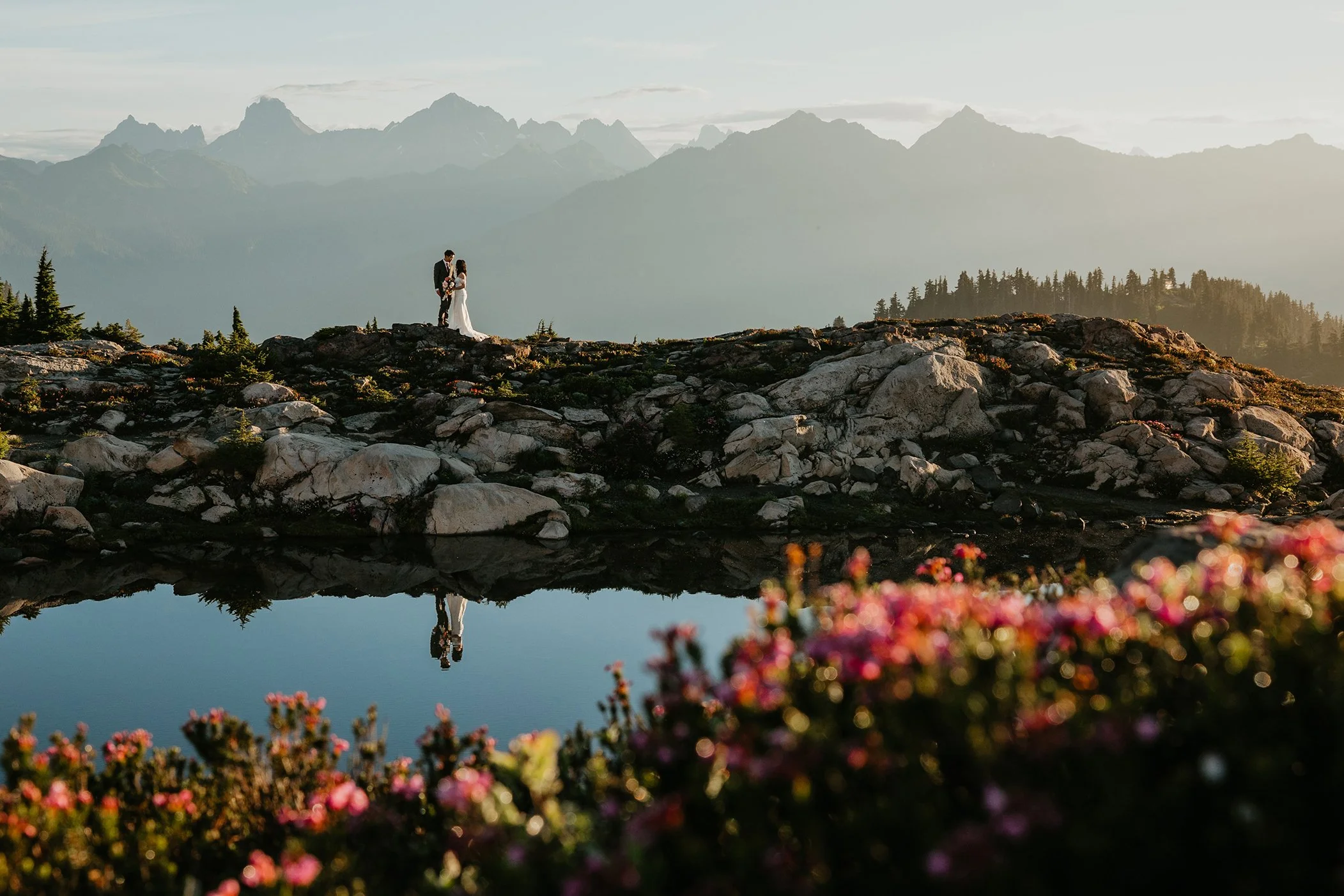 A couple stands on rocky terrain near a small lake with mountains in the background, illuminated by soft, natural light.