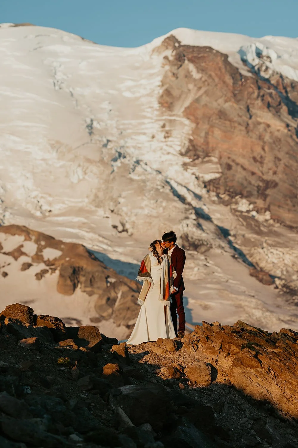 Newlyweds standing together on a rocky ridge as warm light hits the glaciers and rugged mountain terrain behind them