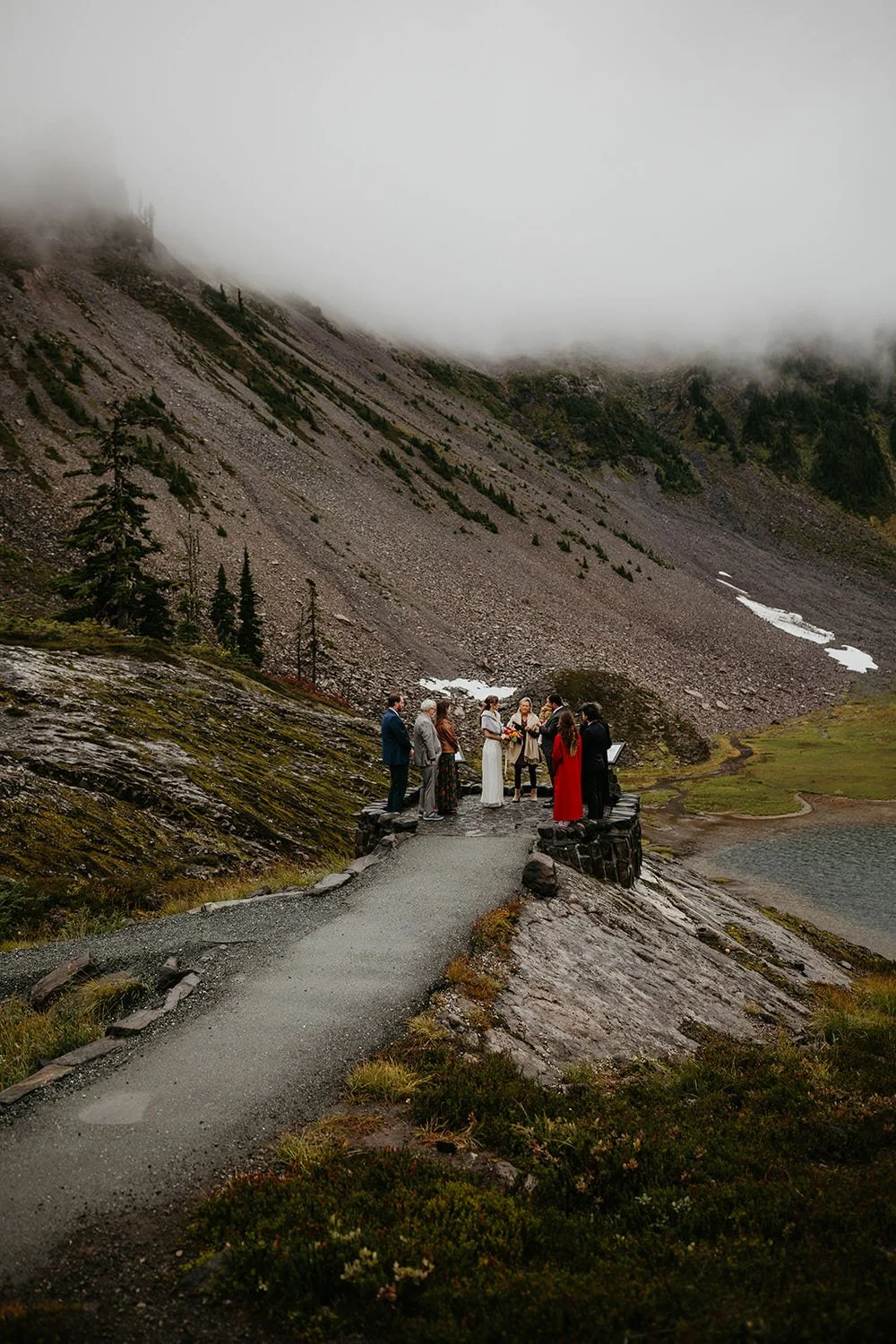 A small group gathers for a mountain elopement ceremony along a foggy alpine trail, surrounded by steep rocky slopes, wild terrain, and low clouds.